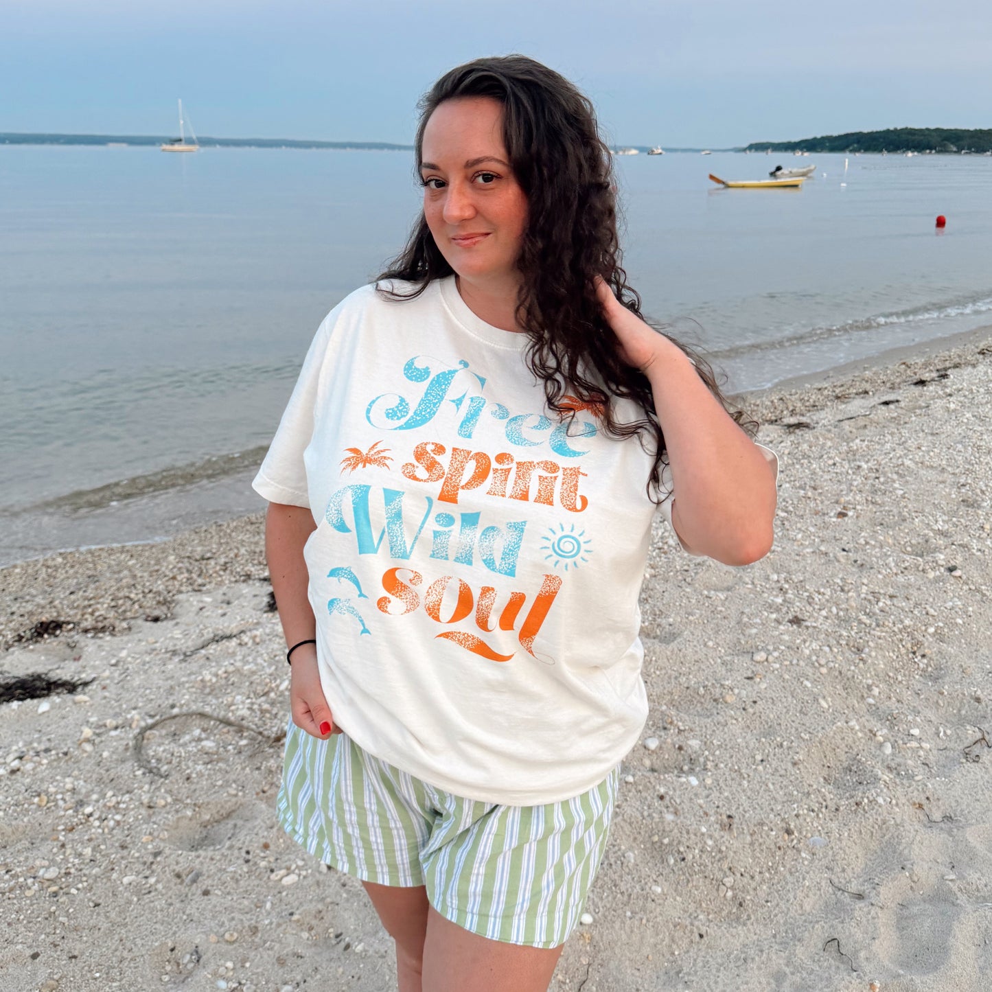 Woman standing on a beach wearing a white t-shirt with colorful text and striped shorts.