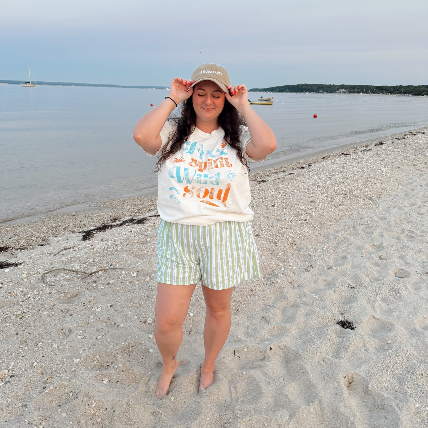 Woman standing on a sandy beach wearing a white t-shirt with colorful text and green shorts.