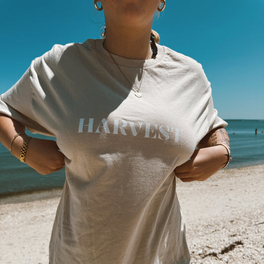 Person wearing a beige t-shirt with 'HARVEST' text on a beach.