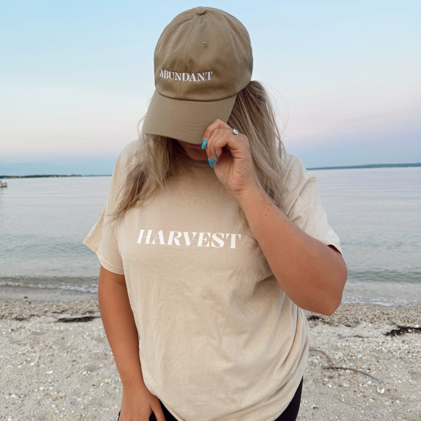 Person wearing a beige 'Harvest' t-shirt and brown cap on a beach