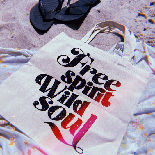 Tote bag with 'Free Spirit Wild Soul' text on sand with flip-flops